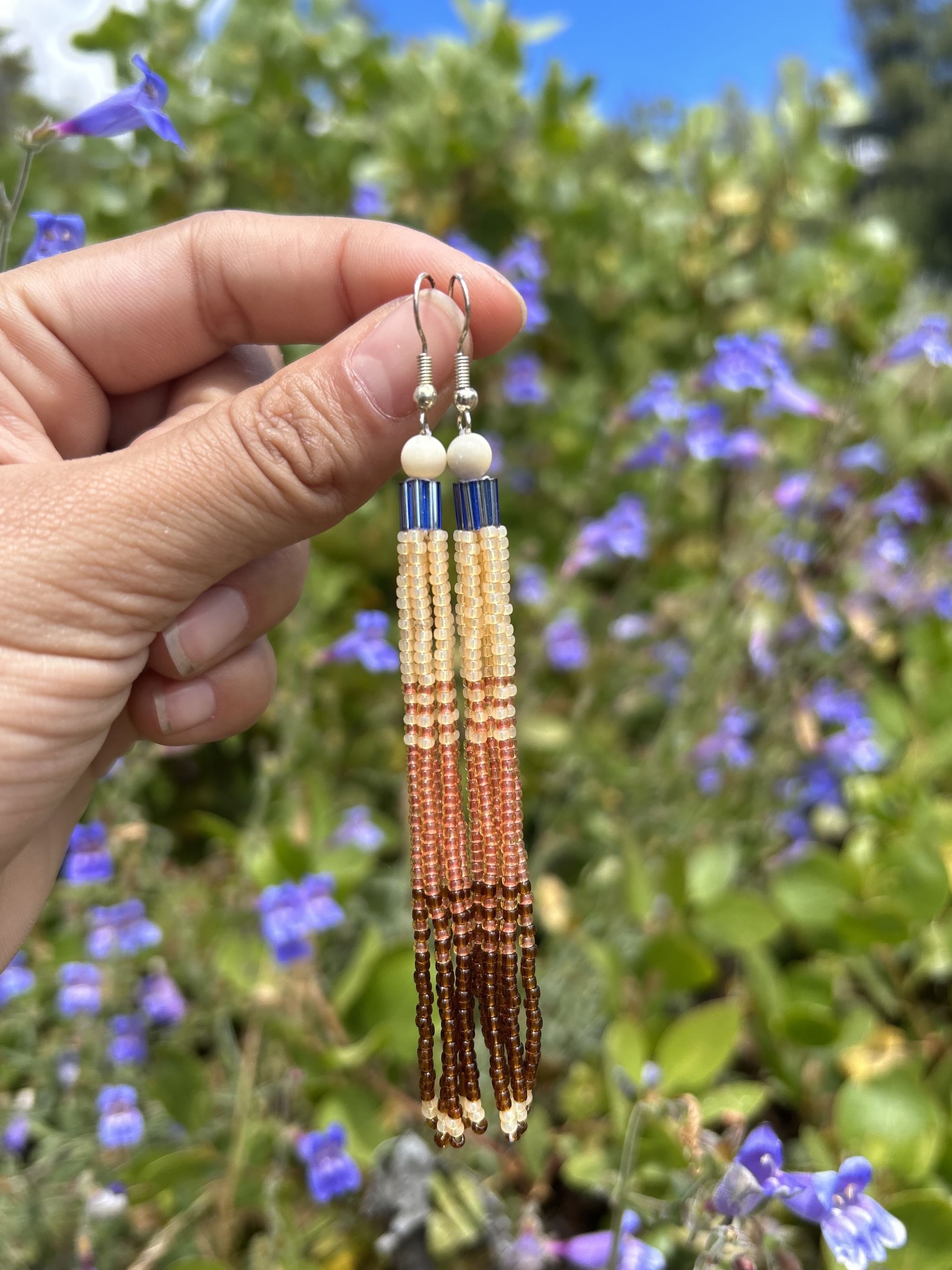 Sunset ombre tassel earrings held against a wildflower garden background