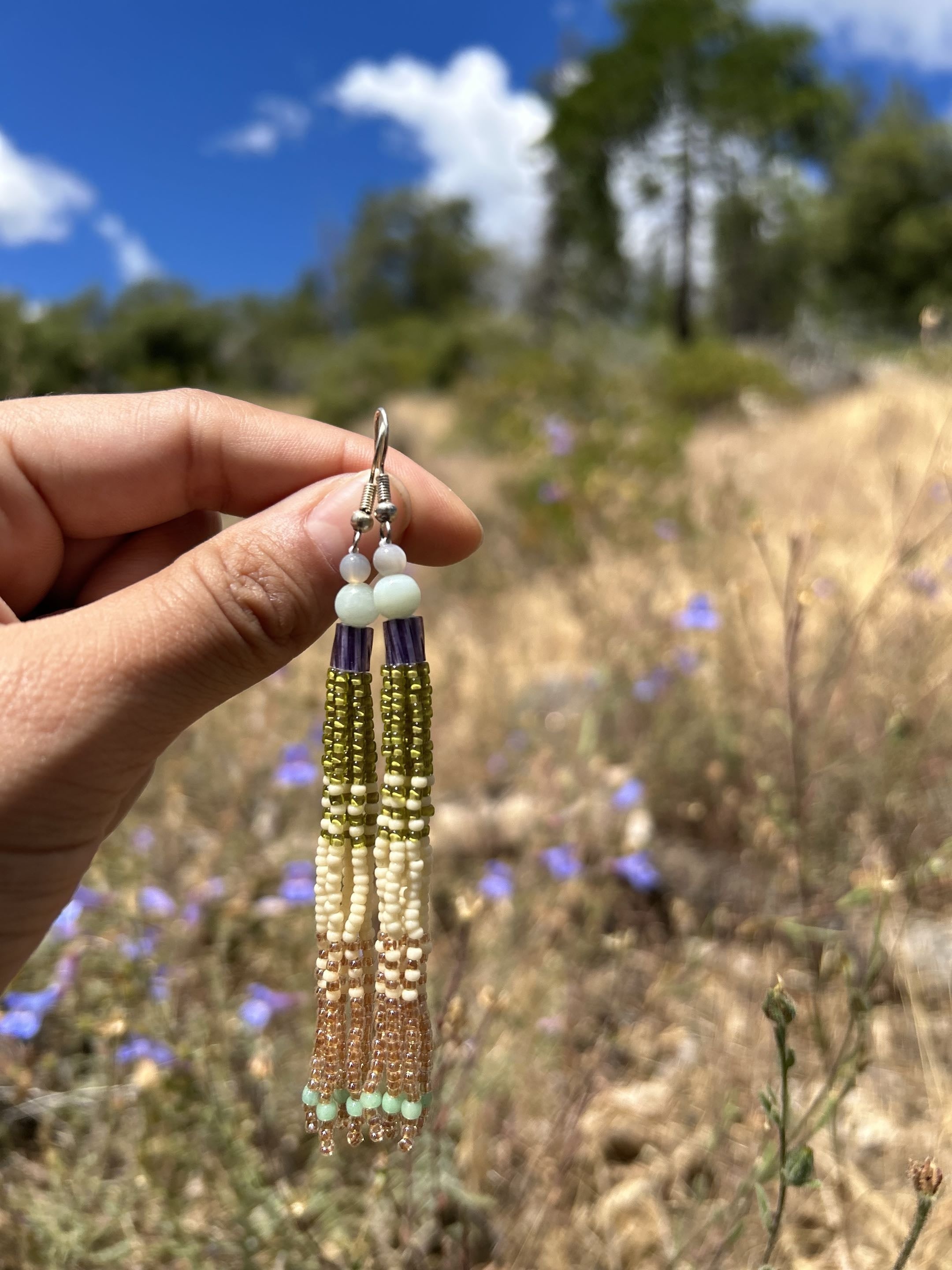 Desert sky tassel earrings held against an open wildflower field under blue sky
